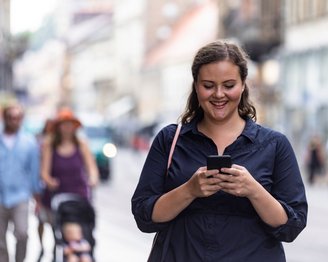 Eine Frau in einem dunkelblauen Hemd steht auf einer belebten Straße und schaut auf ihr Smartphone. Im Hintergrund sind weitere Passanten und Geschäfte zu sehen.