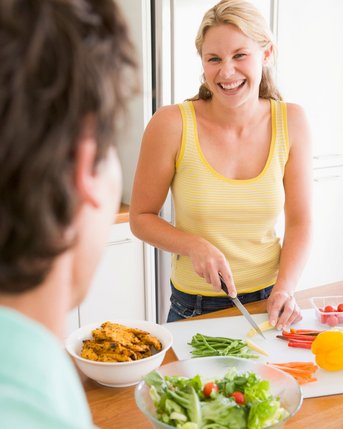 Eine Frau schneidet Gemüse in einer modernen Küche, während sie einem Mann gegenübersteht, der sie ansieht. Eine Schüssel mit Salat und eine andere mit Essen sind ebenfalls sichtbar.