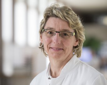 Eine Frau mit blonden, lockigen Haaren und Brille trägt einen weißen Kittel. Sie lächelt freundlich und steht in einem hellen, modernen Innenraum.