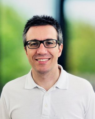 Ein Mann mit Brille und kurzem, dunklem Haar trägt ein weißes Polohemd und lächelt vor einem unscharfen, grünen Hintergrund.