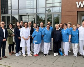 Eine Gruppe von 18 Personen in medizinischer Kleidung steht vor einem Gebäude mit dem Schriftzug "Wald-". Sie lächeln und posieren gemeinsam.