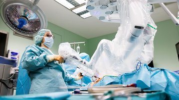 Ein Chirurg in Schutzkleidung bedient ein robotergestütztes Operationssystem in einem modernen Operationssaal. Der Patient ist auf dem Operationstisch positioniert, umgeben von medizinischen Instrumenten und Geräten.