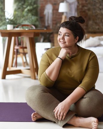 Eine Person sitzt auf einer Yogamatte, trägt ein olivgrünes Oberteil und graue Leggings. Sie hat die Beine gekreuzt und schaut nachdenklich zur Seite, während sie in einem hellen Raum mit natürlichem Licht sitzt.