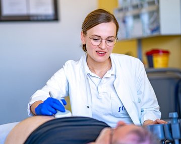 Eine Ärztin führt eine Untersuchung bei einem Patienten durch, der auf einer Liege liegt. Sie verwendet ein medizinisches Gerät zur Diagnose.