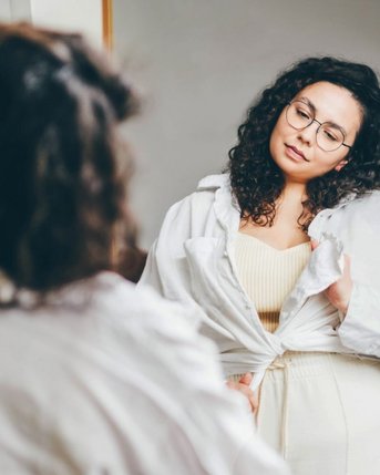 Eine Frau mit lockigem Haar und Brille steht vor einem Spiegel, trägt eine weiße Bluse und ein helles Oberteil. Sie schaut nachdenklich auf ihr Spiegelbild und hält die Bluse leicht geöffnet.