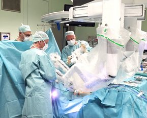 Ein chirurgisches Team in einem Operationssaal führt einen Eingriff durch. Die Ärzte tragen Schutzkleidung und Masken, während sie sich auf den Patienten konzentrieren, der von blauen Tüchern umgeben ist.