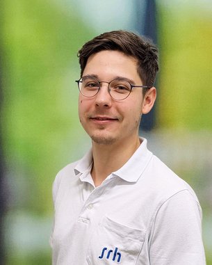 Junger Mann mit Brille und kurzem, dunklem Haar, trägt ein weißes Polohemd mit einem blauen Logo. Hintergrund unscharf.