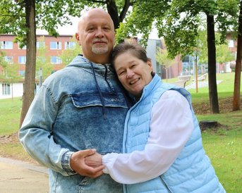 Ein älteres Paar steht im Freien, umarmt sich und lächelt. Der Mann trägt eine Jeansjacke, während die Frau eine hellblaue Weste über einem weißen Oberteil trägt. Bäume und Gebäude sind im Hintergrund sichtbar.