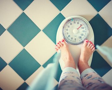 Eine Person steht barfuß auf einer digitalen Waage, die auf einem schachbrettartigen Boden platziert ist. Die Zeiger der Waage zeigen auf ein Gewicht.