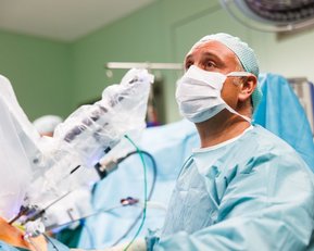 Ein Chirurg in OP-Kleidung und einer Gesichtsmaske beobachtet einen chirurgischen Eingriff. Er trägt eine Haube und ist in einem Operationssaal mit medizinischen Geräten und sterilen Tüchern umgeben.