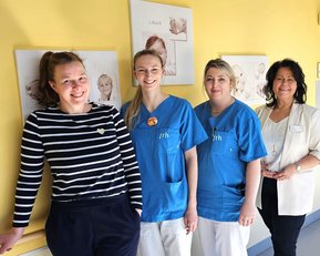 Vier Frauen stehen in einem Flur, gekleidet in medizinische Kleidung. Zwei tragen blaue Oberteile, eine trägt ein gestreiftes Oberteil, und eine weitere trägt eine weiße Jacke. Im Hintergrund sind Zeichnungen von Kindern an der Wand.