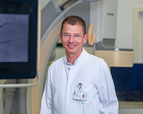 Ein Arzt in einem weißen Kittel steht in einem modernen medizinischen Raum mit einem großen Bildschirm im Hintergrund.