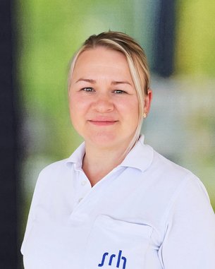 Eine Person mit blonden Haaren trägt ein weißes Polohemd mit dem Logo "srh". Sie lächelt und steht vor einer unscharfen, hellen Hintergrundszene.