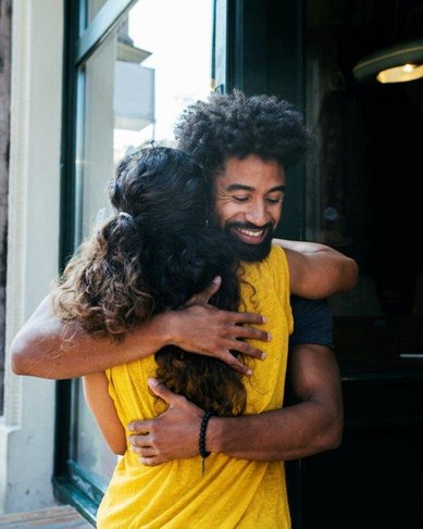 Ein Mann und eine Frau umarmen sich vor einem Gebäude mit großen Fenstern. Der Mann hat lockiges Haar und trägt ein dunkles T-Shirt, während die Frau ein gelbes Oberteil trägt und lange, lockige Haare hat.