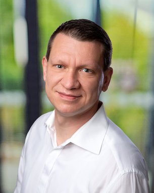 Ein Mann mit kurzen, braunen Haaren und einem weißen Hemd lächelt in die Kamera, vor einem unscharfen, hellen Hintergrund.