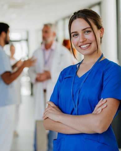 Eine junge Frau in blauer Arbeitskleidung lächelt und steht mit verschränkten Armen im Vordergrund. Im Hintergrund sind mehrere medizinische Fachkräfte in einem Krankenhausumfeld zu sehen, die miteinander sprechen.