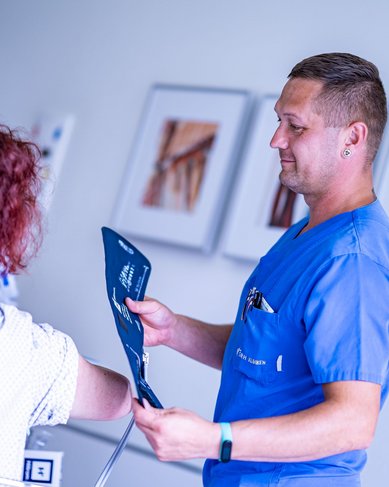Ein Arzt in blauer Arbeitskleidung hält eine Röntgenaufnahme und spricht mit einer Patientin, die auf einem Stuhl sitzt. Im Hintergrund sind Bilder an der Wand zu sehen.