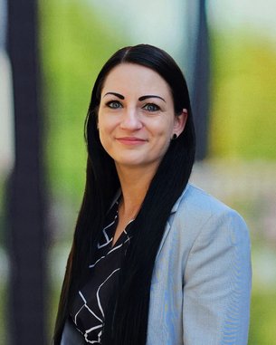 Eine Frau mit langen, schwarzen Haaren trägt einen grauen Blazer und ein schwarzes Oberteil mit Muster. Sie steht lächelnd vor einer unscharfen, hellen Hintergrundlandschaft.