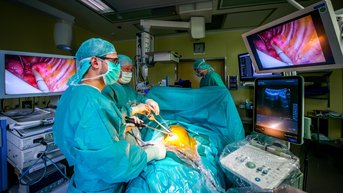 Chirurgisches Team in grüner Kleidung führt einen Eingriff durch, während Bildschirme den Operationsbereich zeigen.