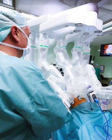 Chirurgisches Team in einem Operationssaal, das ein robotergestütztes chirurgisches System bedient. Die Ärzte tragen OP-Kleidung und Schutzmasken, während ein Monitor im Hintergrund sichtbar ist.