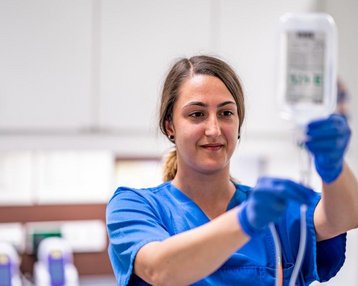 Eine Krankenschwester in blauer Arbeitskleidung hält eine Infusionsflasche in der Hand. Sie trägt Handschuhe und konzentriert sich auf die Vorbereitung der Infusion. Im Hintergrund sind medizinische Geräte sichtbar.