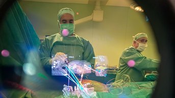 Ein Chirurg in OP-Kleidung arbeitet an einem Patienten, sichtbar durch ein chirurgisches Instrument. Ein zweiter Chirurg ist ebenfalls im Hintergrund zu sehen.