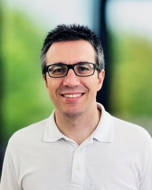 Ein Mann mit Brille und kurzem, dunklem Haar trägt ein weißes Polohemd und lächelt vor einem unscharfen, grünen Hintergrund.