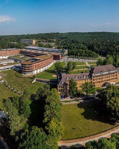 Eine Luftaufnahme eines großen Komplexes mit modernen und historischen Gebäuden, umgeben von Bäumen und Grünflächen. Die Anlage liegt in einer hügeligen Landschaft, die von Wäldern und Feldern geprägt ist.