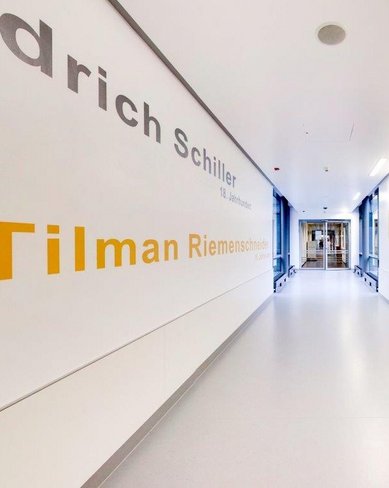 Ein heller Flur mit weißen Wänden, an denen die Namen "Friedrich Schiller" und "Tilman Riemenschneider" in verschiedenen Schriftarten angebracht sind. Am Ende des Flurs sind Türen sichtbar.