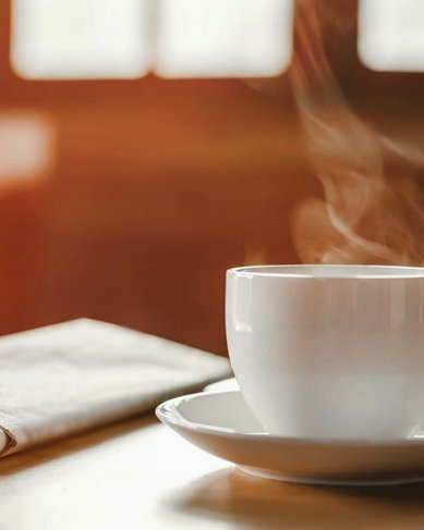 Eine dampfende Tasse Kaffee steht auf einer Untertasse, neben einem gefalteten Stoffserviette. Der Hintergrund zeigt unscharfe Fenster, die warmes Licht hereinlassen.