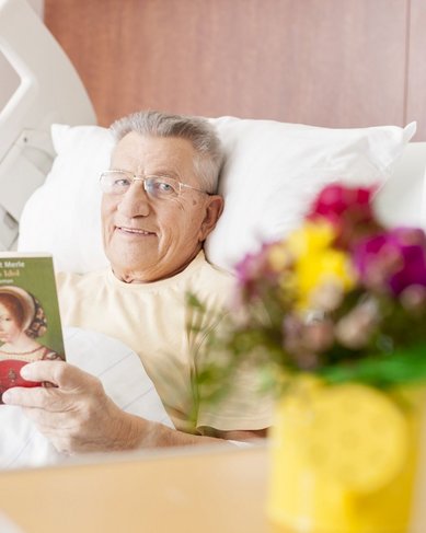 Ein älterer Mann im Krankenhausbett liest ein Buch. Er trägt eine Brille und lächelt. Auf dem Nachttisch steht eine blühende Pflanze in einem gelben Topf.