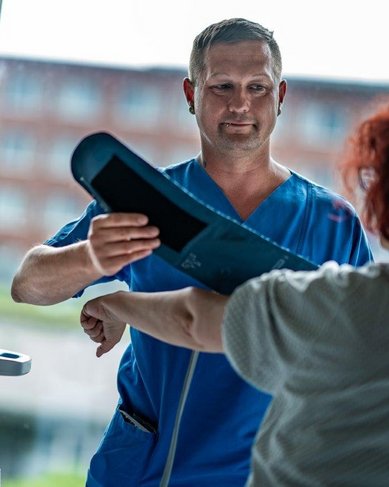 Ein Gesundheitsdienstleister misst den Blutdruck einer Patientin mit einem Blutdruckmessgerät. Der Fachmann trägt einen blauen Kittel und steht in einem hellen Raum mit großen Fenstern. Die Patientin hat lockiges, rotes Haar.