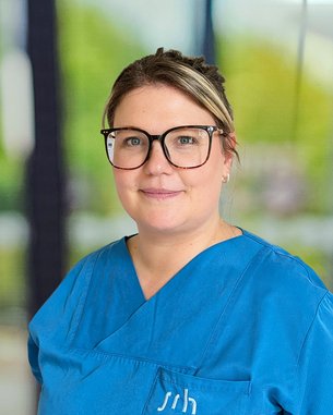 Eine Frau mit Brille trägt ein blaues Oberteil und lächelt. Der Hintergrund zeigt unscharfe grüne und graue Elemente.