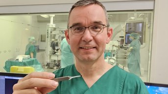 Ein Arzt in grüner OP-Kleidung hält eine Pinzette in der Hand. Im Hintergrund sind medizinisches Personal in OP-Kleidung und ein Operationssaal zu sehen.