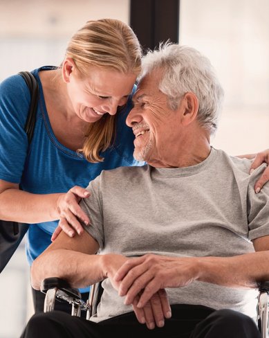 Eine Frau steht hinter einem älteren Mann im Rollstuhl und lächelt ihn an. Der Mann trägt ein graues T-Shirt und sieht freundlich aus. Die Szene vermittelt eine Verbindung zwischen den beiden.