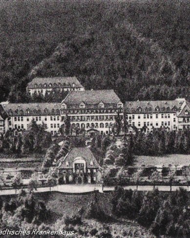Städtisches Krankenhaus in Gera, umgeben von Bäumen und Grünflächen. Das Gebäude zeigt eine historische Architektur mit mehreren Flügeln und einem zentralen Eingang. Ein Schornstein ist ebenfalls sichtbar.