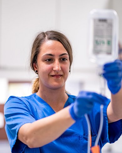 Eine Krankenschwester in blauer Arbeitskleidung hält eine Infusionsflasche in der Hand und bereitet sie vor. Sie trägt Handschuhe und steht in einem modernen medizinischen Raum.
