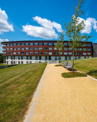 Ein gepflasterter Weg führt durch eine grüne Wiese zu einem modernen, mehrstöckigen Gebäude mit großen Fenstern. Der Himmel ist blau mit einigen Wolken, und es gibt Bäume in der Umgebung.