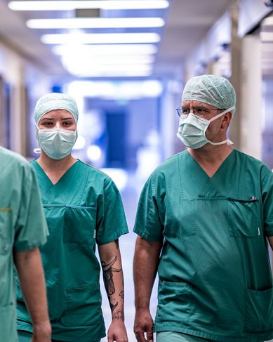 Drei medizinische Fachkräfte in grünen OP-Kitteln und Mundschutz gehen durch einen Krankenhausflur. Sie tragen Operationsmützen und haben einen konzentrierten Ausdruck.