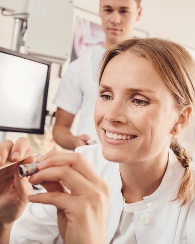 Eine Ärztin untersucht das Maul eines Patienten mit einem medizinischen Instrument, während ein weiterer Mitarbeiter im Hintergrund beobachtet. Beide tragen medizinische Kleidung in einer klinischen Umgebung.